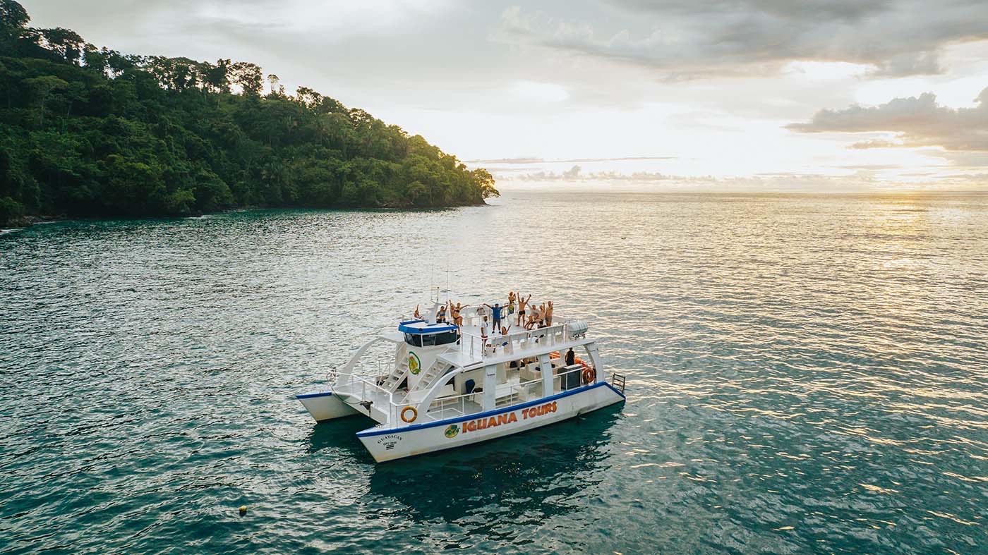 The Iguana Tours catamaran off a forested headland near Manuel Antonio at sunset, with guests waving from the top deck