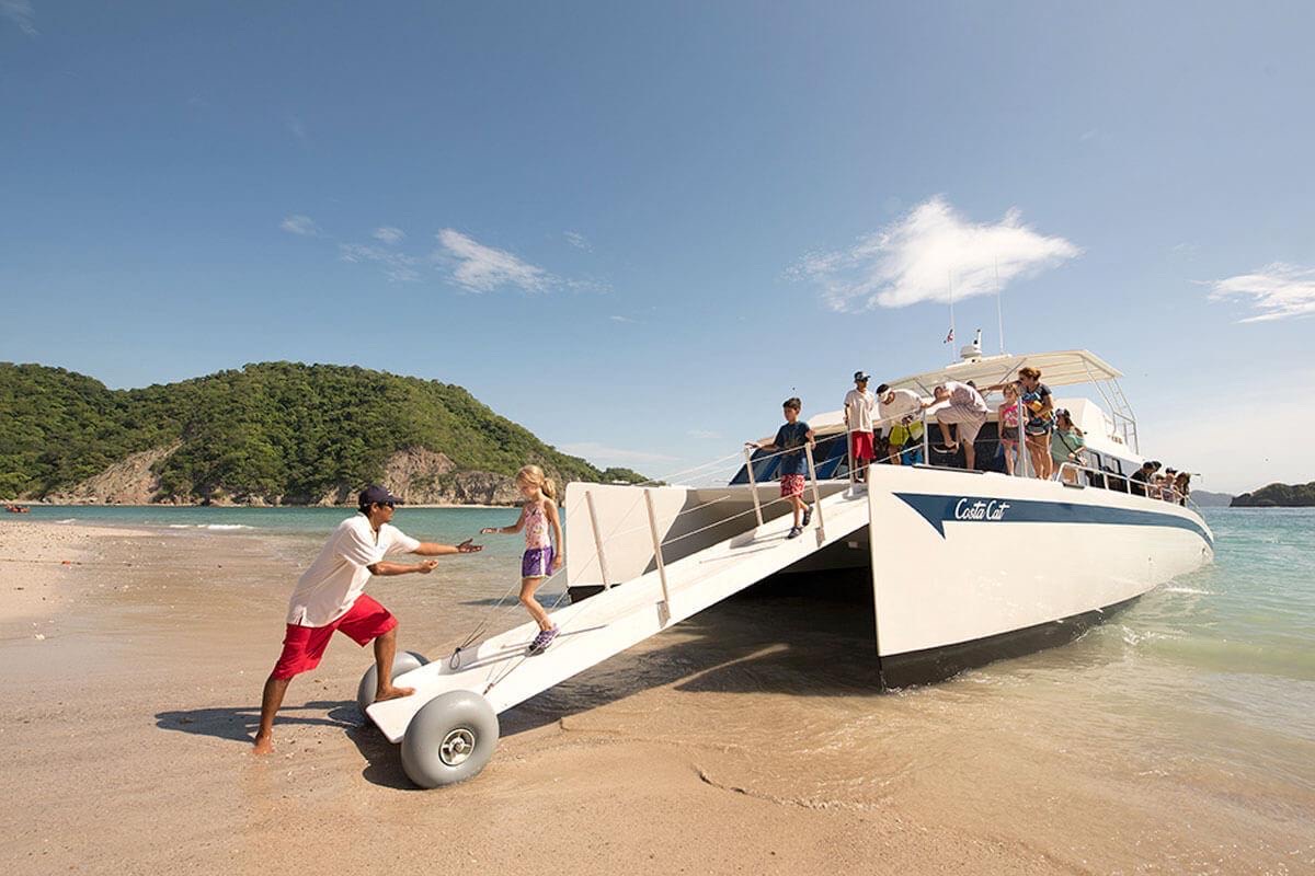 A crew member helps a child down the wheeled boarding ramp of a catamaran pulled up on the beach at Tortuga Island, on a JacoVIP day trip