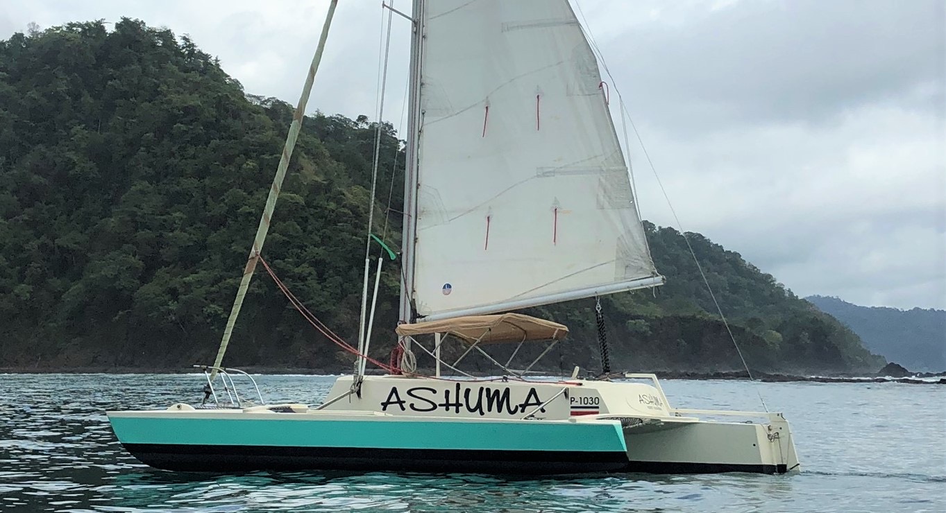 Kayak Jaco's 25-foot Searunner trimaran Ashuma under sail off a green headland on the Central Pacific coast