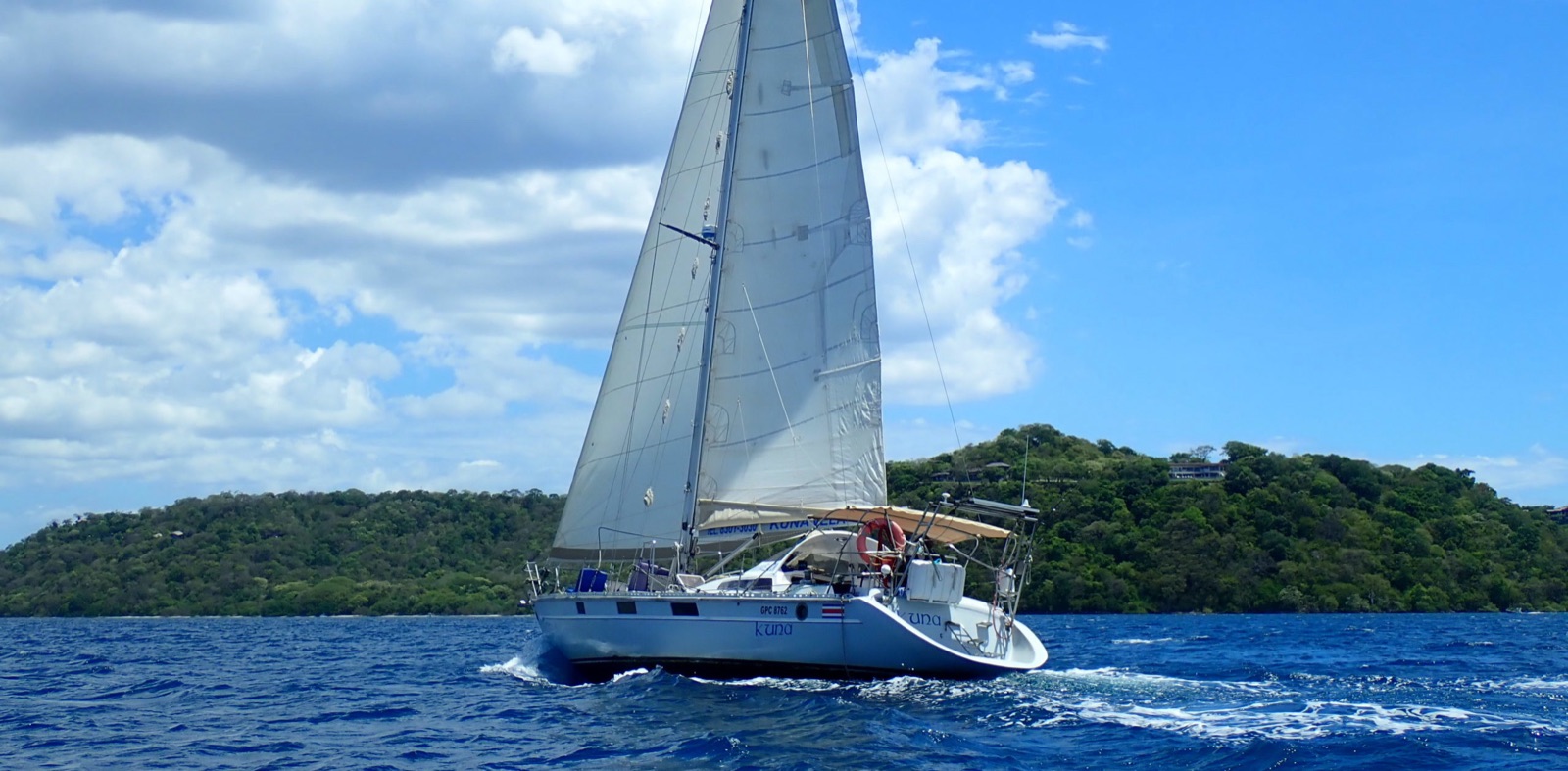 Kuna Vela's white sloop Kuna under full sail on blue water, with a green, forested shoreline behind it