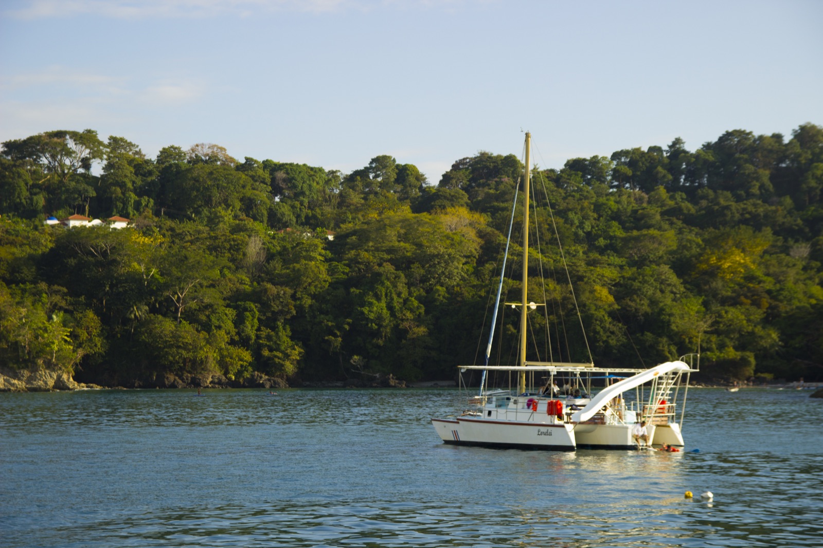 Sunset Sails Tours' boat Lorelei at anchor in a forested bay, with guests on deck, a water slide and a snorkeler at the stern