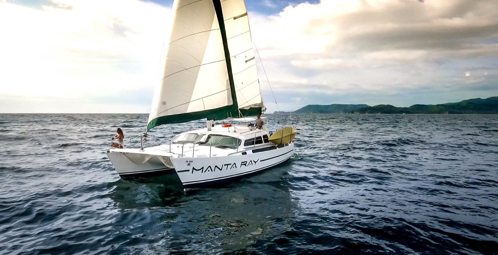 The original Manta Ray catamaran under sail off the Guanacaste coast, its name painted on the hull and a paddleboard strapped to the side