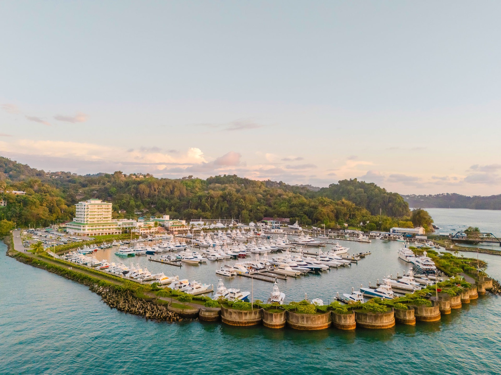 Aerial view of Marina Pez Vela in Quepos at sunset, showing the full basin of docked boats ringed by a round-pillared breakwater, with green hills behind