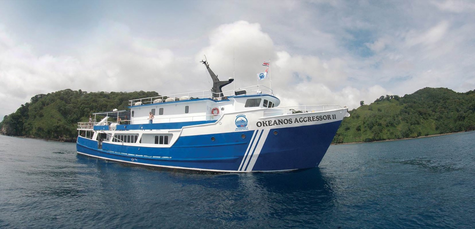 The blue-and-white dive yacht Okeanos Aggressor II at anchor off a forested island shore