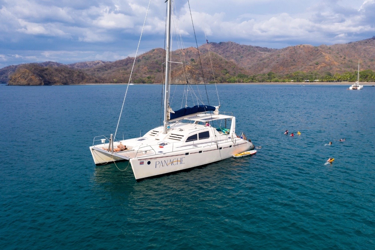 The catamaran Panache at anchor in a turquoise bay with snorkelers and a paddleboard in the water and dry hills behind