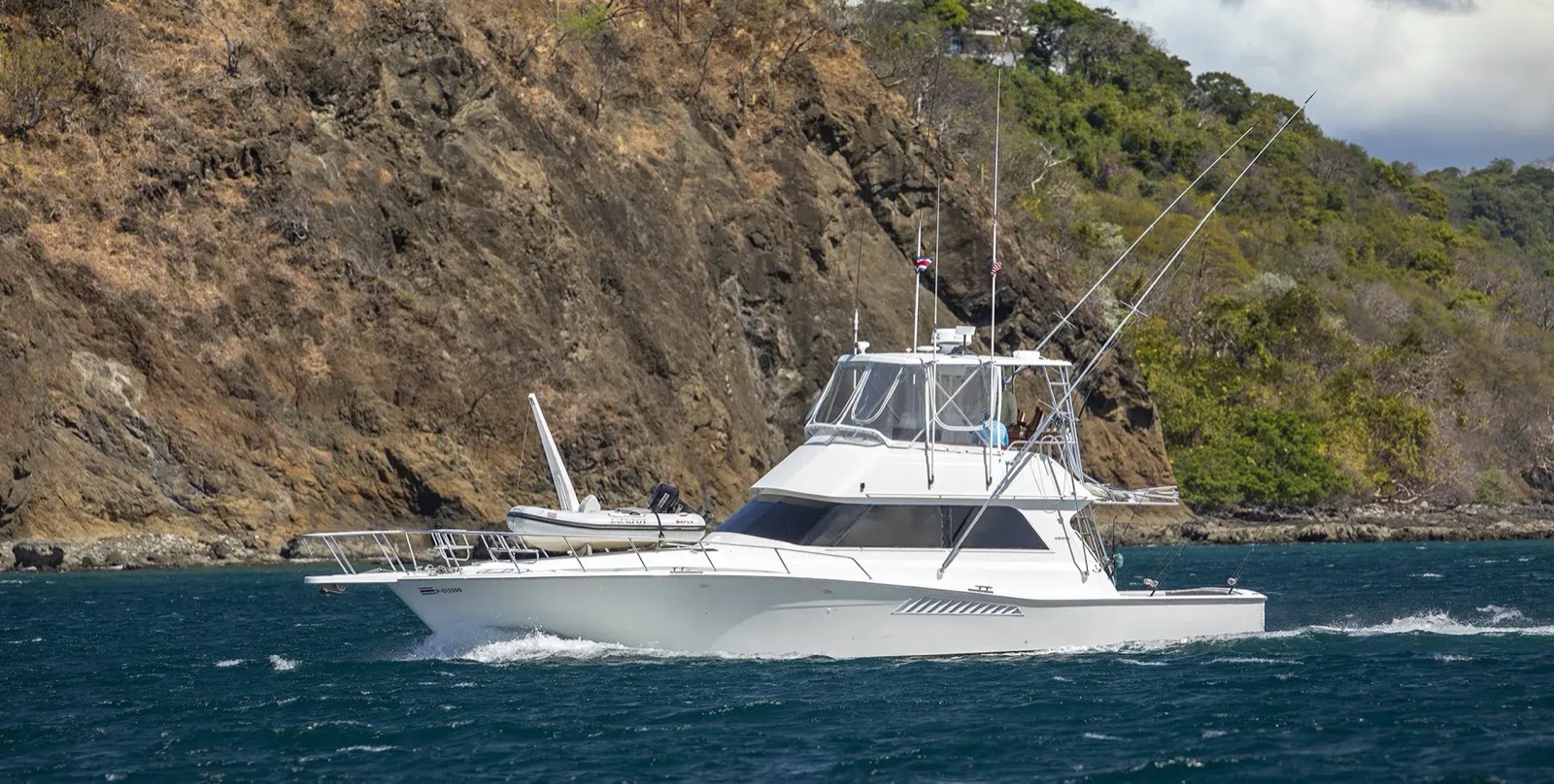 A Papagayo Sport Fishing Charters sportfishing boat with outriggers up, cruising past a rocky, forested headland