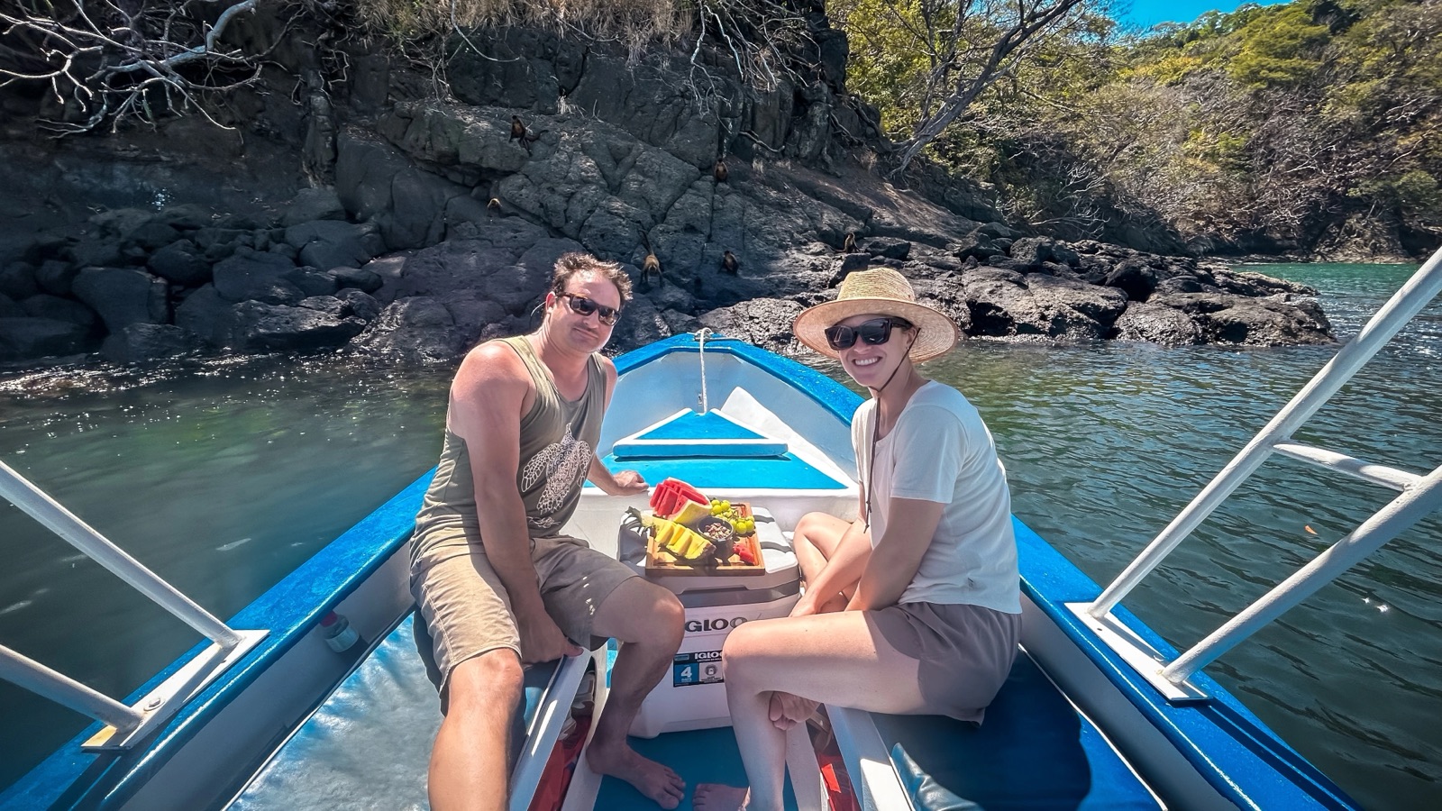 Two guests sharing a fruit plate in the bow of a Pochote Ocean Adventures boat, anchored by a rocky shoreline