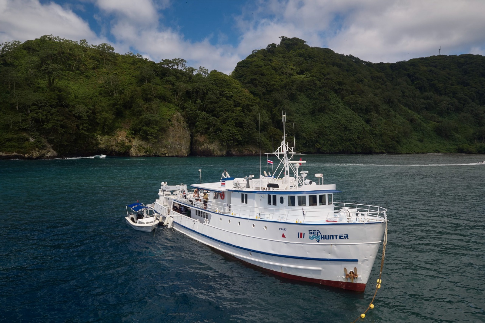 Undersea Hunter's white liveaboard Sea Hunter at anchor below the forested cliffs of Cocos Island, with a dive skiff tied alongside