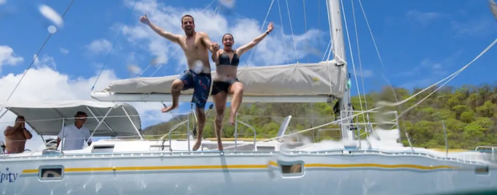 Two guests leaping off the side deck of the sailing yacht Serendipity while a crew member watches from under the cockpit shade